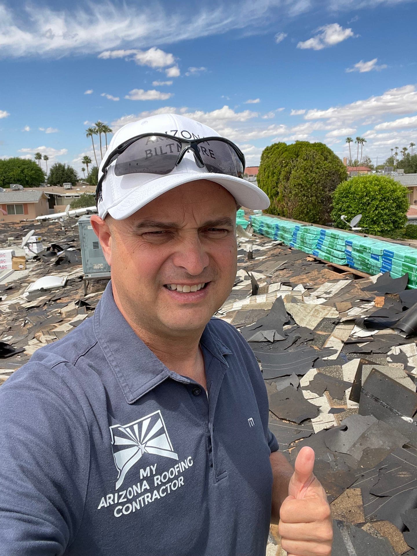 James Naylor, owner of My Arizona Roofing Contractor, on a roof during a tear-off — company polo, thumbs up, stripped decking and stacked new material behind him