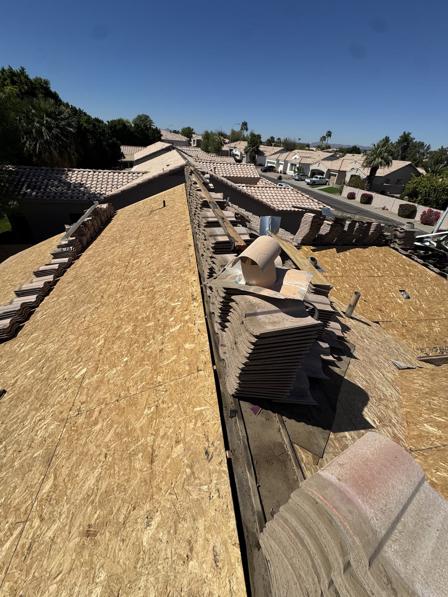 Looking along the ridge of a tile roof during an underlayment replacement — one slope stripped to new decking, the other still carrying stacked tile, above a street of tile-roofed Phoenix homes.