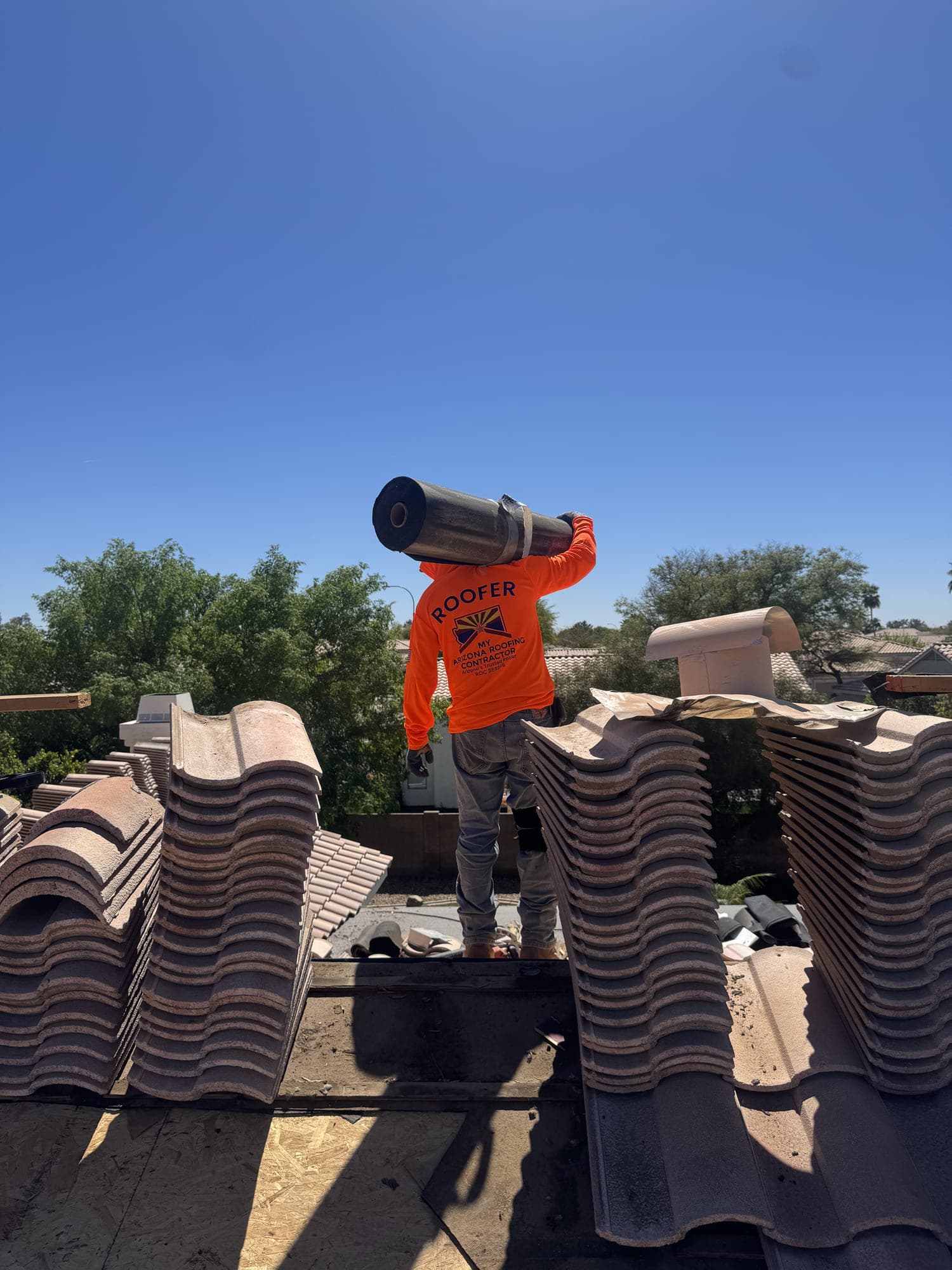 A roofer carrying a roll of underlayment across a roof stripped back to new decking, with concrete tile lifted and stacked in rows along the slope beside him.