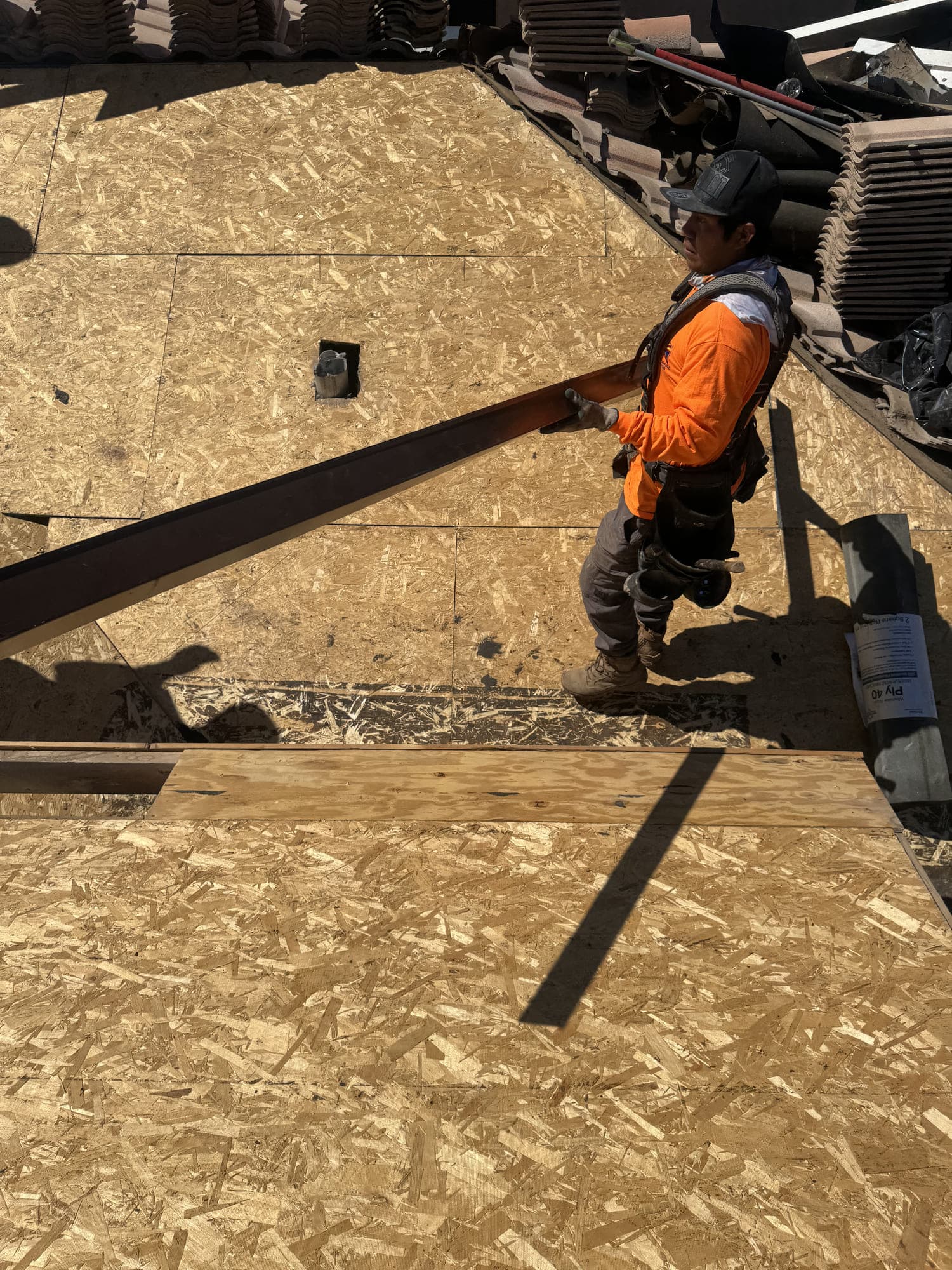 A roofer carrying a length of timber across newly laid roof decking, with rolls of underlayment and stacks of lifted concrete tile waiting at the ridge behind him.