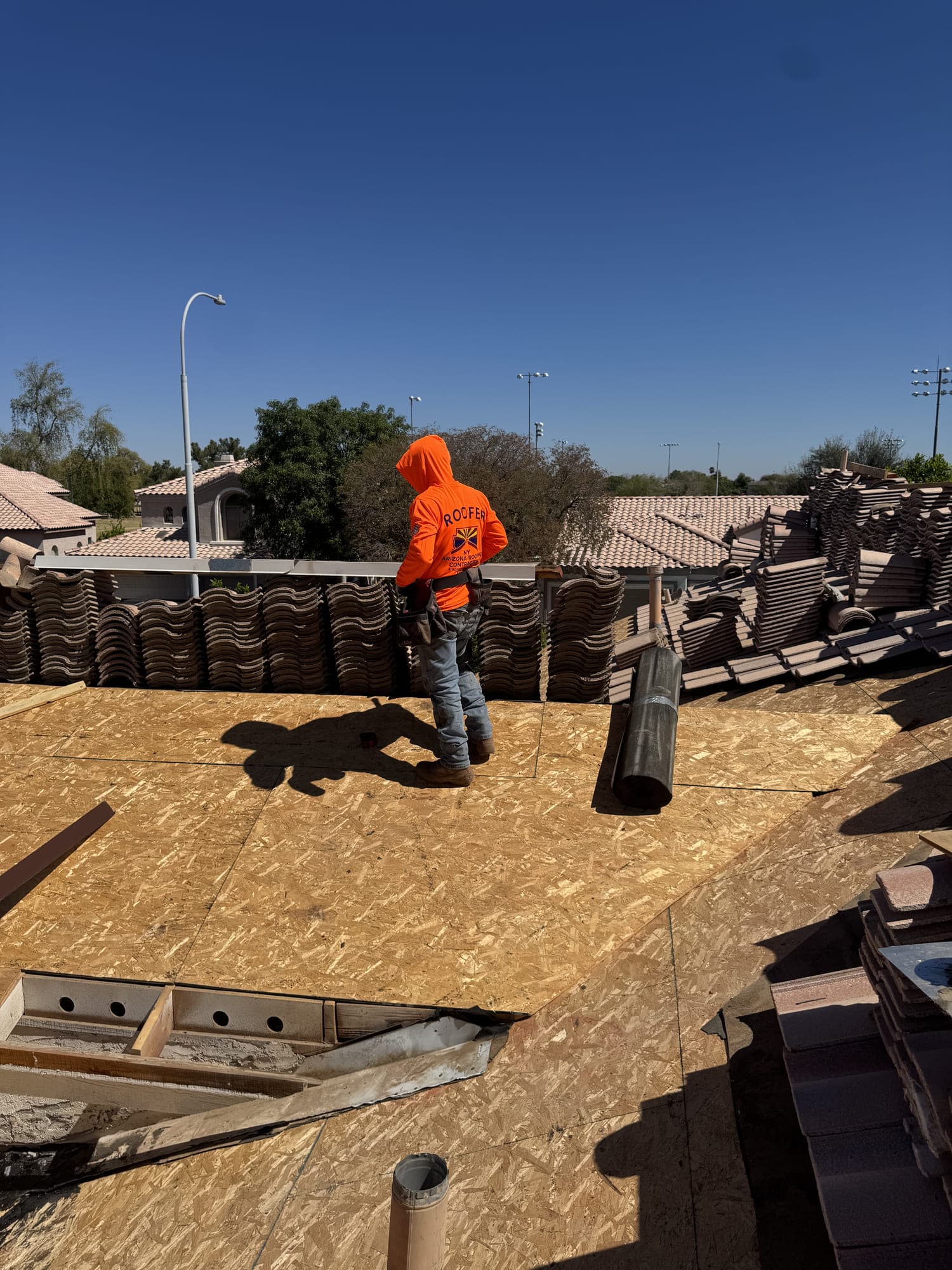 A roofer standing on newly laid decking beside long rows of concrete tile lifted from the roof, with damaged decking cut away and open rafters in the foreground.
