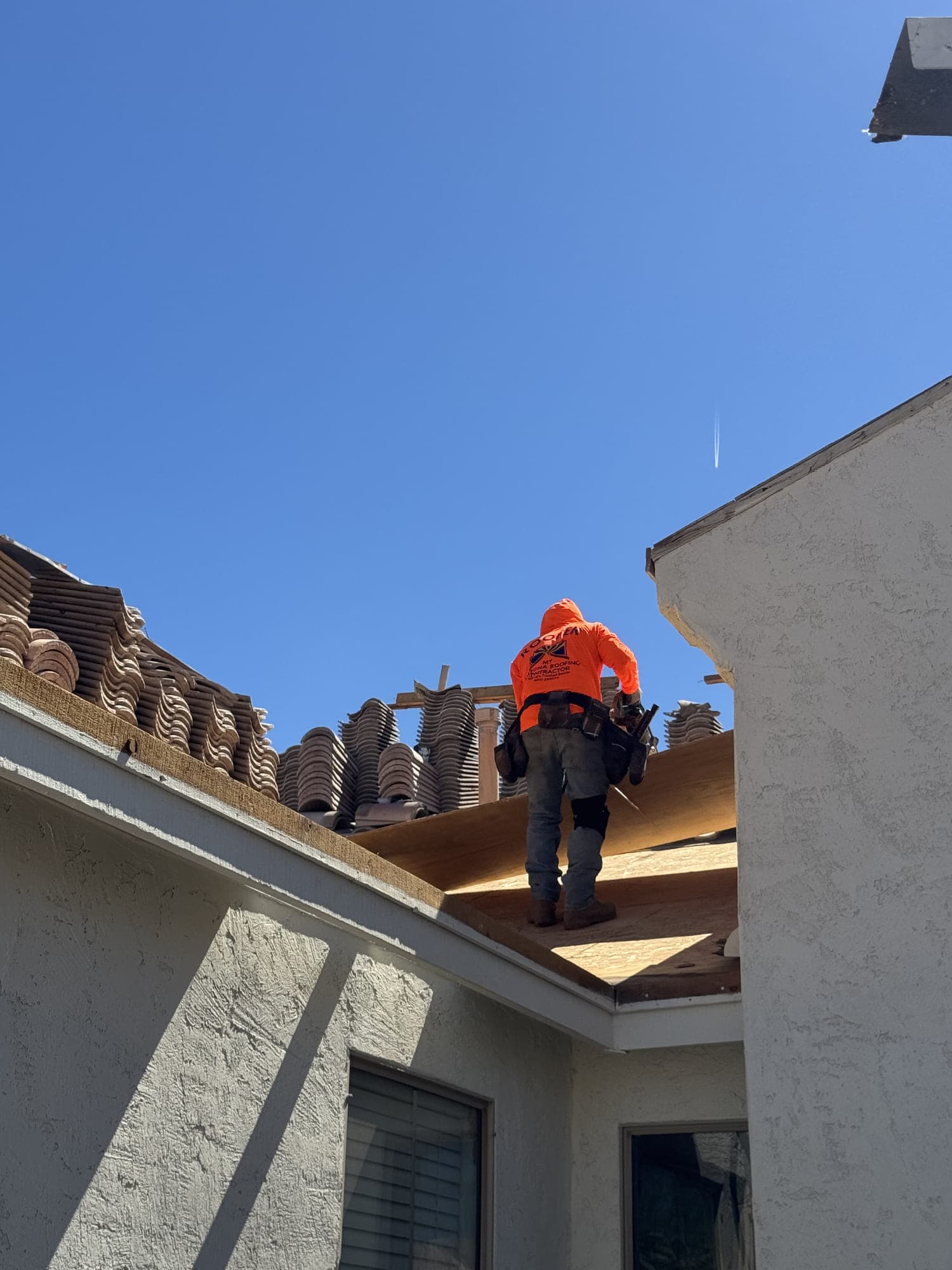A roofer standing on newly laid decking beside a stucco wall, with long rows of concrete tile stacked along the slope above him.