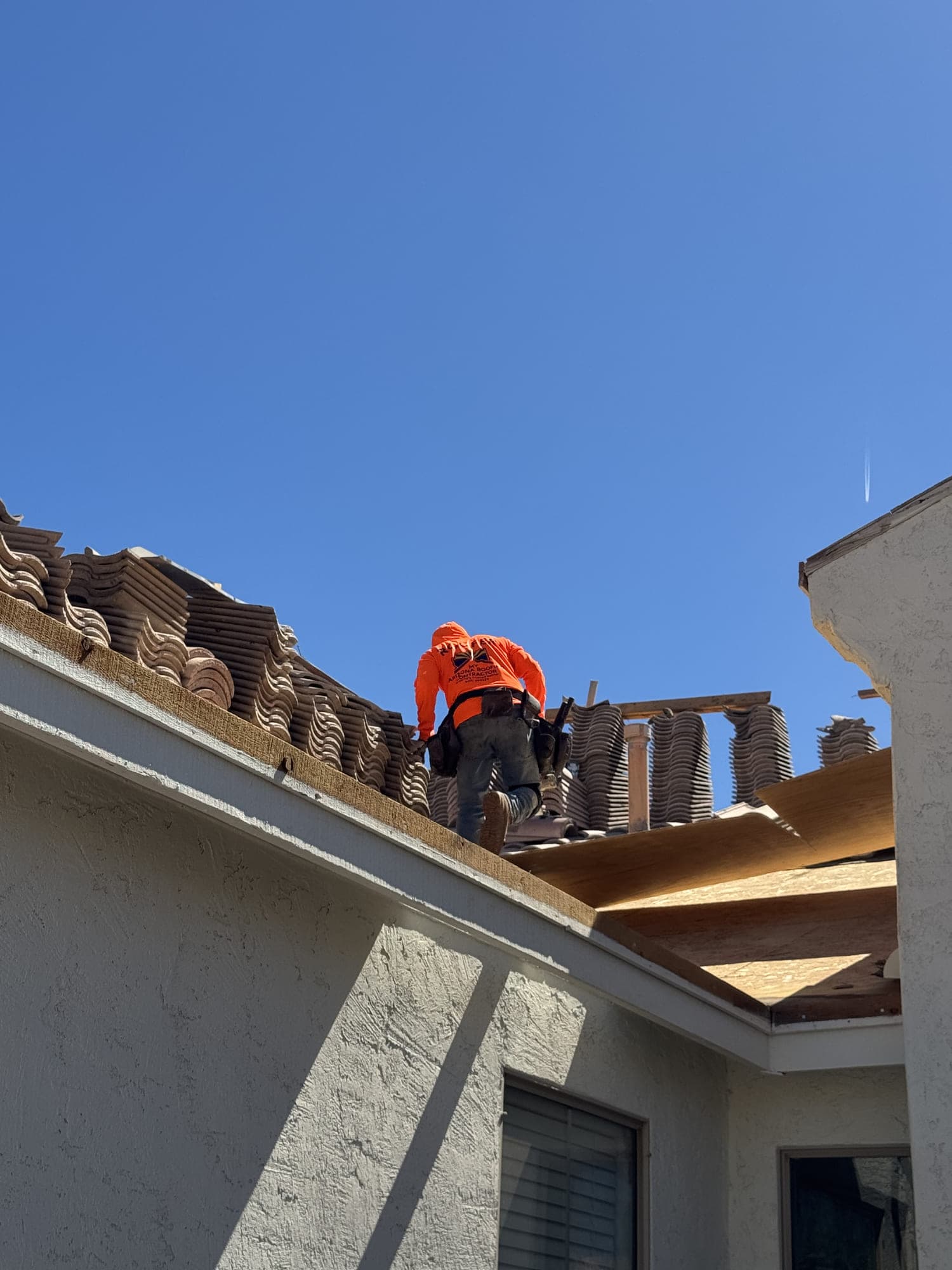 A roofer working along a course of concrete tile near the ridge, with lifted tile stacked in rows behind him and new decking exposed on the low-slope section alongside.
