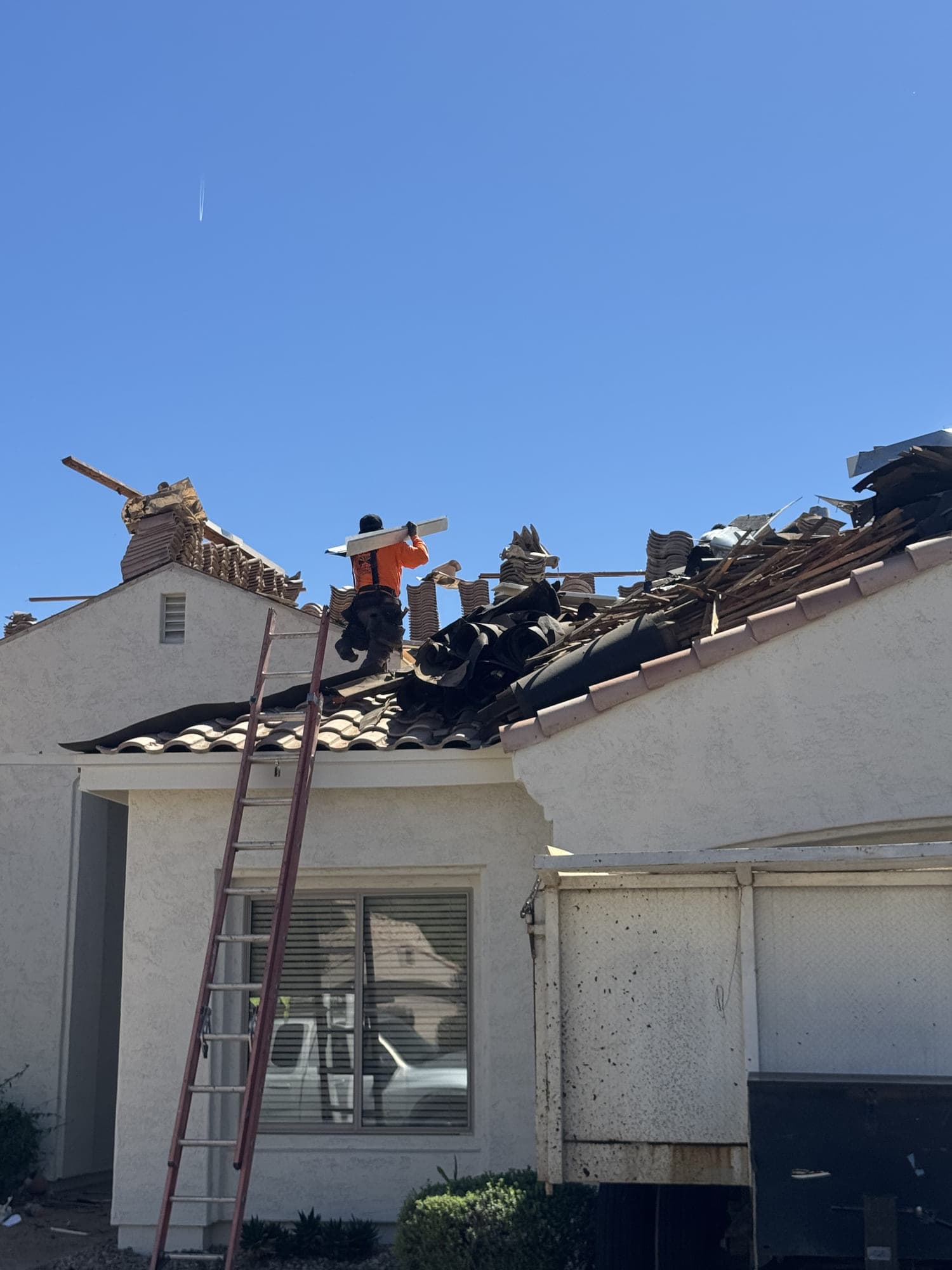 A tile roof part way through a tear-off seen from the driveway, old underlayment stripped back in rolls, tile and battens stacked along the ridge, and a ladder up to the eave.